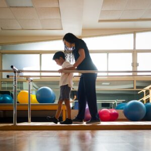 A physio helps a young boy with walking excercises in a gymnasium